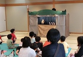 人形劇「おむすびころりん」を公演中。おじいさんがネズミの屋敷でネズミたちに迎えられているシーンと、それを見つめる子供たち。