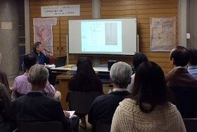 画像:古文書を投影しながら話をされる講師。壁には2枚の絵図も掲示されている。