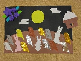 画像:満月の夜、桔梗の花と月見団子が供えられたすすきの原っぱにウサギが集まっています。