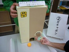 写真：長方形のガチャポン箱を操作して、中からピンポン玉が転がり出たところ。