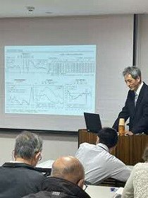 写真 ：スクリーンの前に立って話をする講師と話を聞く参加者たち。