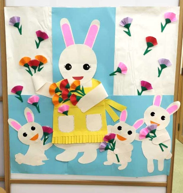 エプロンを付けたうさぎに、3匹のこうさぎがカーネーションを渡しています。5月の壁面飾りです。