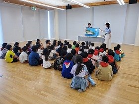 写真　集まって座っている子供たちに、大型絵本『ふねくんのたび』を読んでいる様子