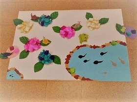 写真　青やピンクのあじさいが咲いて、その周りにはかえるやかたつむりが並んでいる。池の中をおたまじゃくしが泳いでいる。