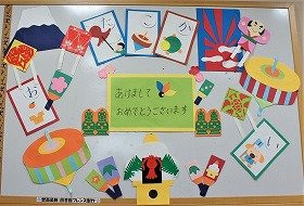 写真　「あけましておめでとうございます」と書かれた四角いメッセージカード。門松。鏡餅。奴凧。羽子板。いろはカルタ。梅の花。コマ。初日の出。富士山。1月の壁面装飾です。