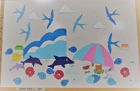 写真　海水浴の風景。空には雲とかもめ。海には大小さまざまな魚、いるか、たこ、くじら、船。砂浜にはパラソル。浮き輪やボールで遊ぶ動物たち。8月の壁面装飾です。