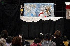 写真4：人形劇「おばあさんのたんじょうび」の写真。舞台中央に雪だるま、その右横におばあさんがいる。舞台の左袖からうさぎ、きつね、くまが顔を出している。
