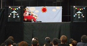写真1：人形劇「のせてのせて」の写真。赤い自動車にうさぎやくまたちが乗っている。舞台の中央には太陽が輝き、両端にはメリーゴーランドがある。