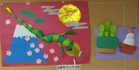 写真赤い画用紙の上に、紙で作った青い富士山と緑の龍、金色のお月様があります。その横には、門松や鏡餅の飾りもあります。