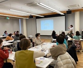 写真1：健康医療情報講座「ハンドケア講習会」で前方のスクリーンを見ながら講師の指示のもとハンドクリームを塗布する参加者の様子。