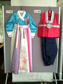女子の韓服、男子の韓服。女子はブルーの上着にピンク、紫、クリームのロングスカート。男子は赤い上着に