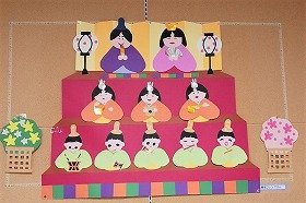 写真　色画用紙で作った雛人形の三段飾りです。一段目はお内裏様、二段目は三人官女、三段目には五人囃子が並んでいます。