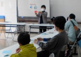折り紙の説明をする先生と、それを聞く参加者。