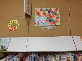 橙、赤、黄、緑のもみじが空色の画用紙いっぱいに広がっています。左下には読みかけの本が開いています。