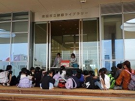 写真：ウッドデッキに集まって座り、大型絵本『しりとりのだいすきなおうさま』の読み聞かせを楽しんでいる子供たち